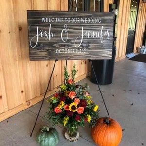 May include: A rustic wooden wedding sign with the text "Welcome to our wedding Josh & Jennifer October 9, 2021" in white lettering. The sign is displayed on a black easel with fall flowers and a pumpkin in the foreground.