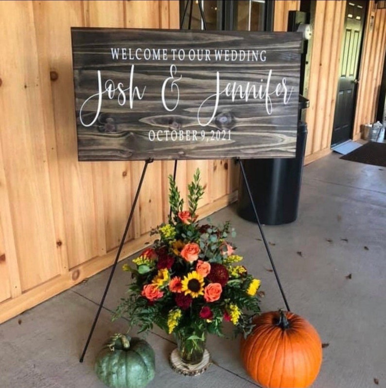 May include: A rustic wooden wedding sign with the text "Welcome to our wedding Josh & Jennifer October 9, 2021" in white lettering. The sign is displayed on a black easel with fall flowers and a pumpkin in the foreground.