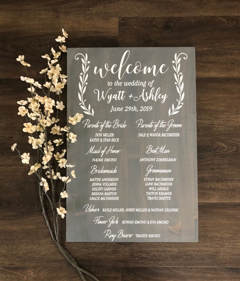 Puede incluir: Un letrero de madera r&uacute;stico con texto blanco anunciando una boda para Wyatt y Ashley el 29 de junio de 2019. El letrero enumera los nombres de los miembros de la fiesta de bodas, incluidos los padres de la novia y el novio, la dama de honor, las damas de honor, el padrino, los padrinos, los acompa&ntilde;antes, las damas de honor y el portador de anillos.