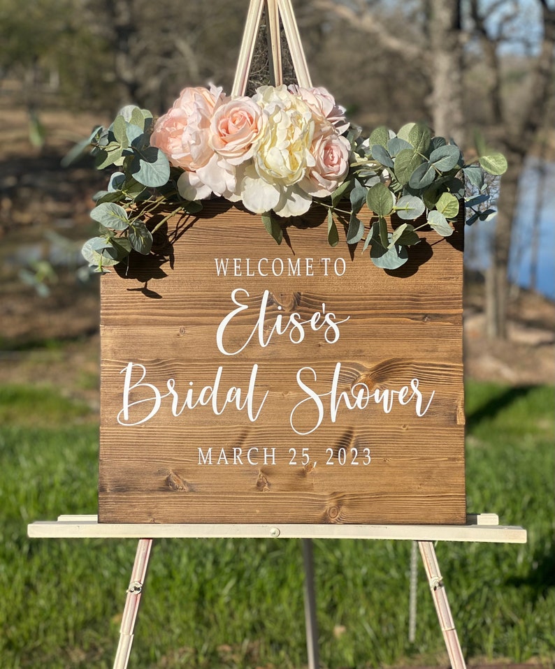May include: A wooden sign with the text "Welcome to Elise's Bridal Shower March 25, 2023" in white script. The sign is decorated with pink and white flowers and eucalyptus leaves, and is displayed on a white easel.