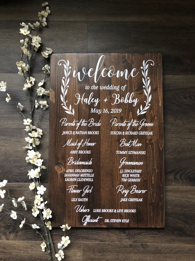 Puede incluir: Un letrero de madera con texto blanco anunciando una boda. El letrero enumera los nombres de la novia y el novio, Haley y Bobby, la fecha de la boda, 16 de mayo de 2019, y los nombres de los miembros de la fiesta de bodas.