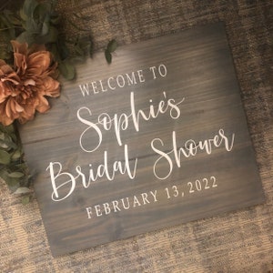 May include: A gray wooden sign with white lettering that reads "Welcome to Sophie's Bridal Shower February 13, 2022." The sign is decorated with a large peach-colored flower and greenery.