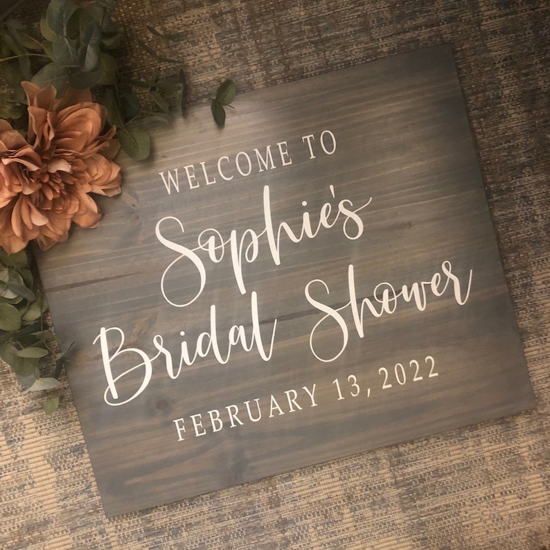 May include: A gray wooden sign with white lettering that reads "Welcome to Sophie's Bridal Shower February 13, 2022." The sign is decorated with a large peach-colored flower and greenery.