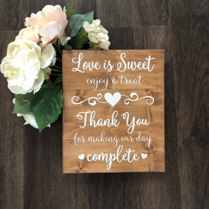 May include: A rustic wooden sign with white text that reads "Love is Sweet enjoy a treat Thank You for making our day complete" with a heart design. The sign is decorated with white hearts and a decorative scroll design.