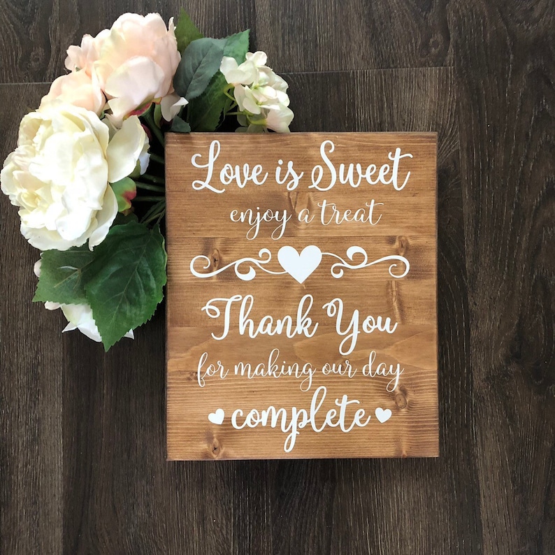 May include: A rustic wooden sign with white text that reads "Love is Sweet enjoy a treat Thank You for making our day complete" with a heart design. The sign is decorated with white hearts and a decorative scroll design.