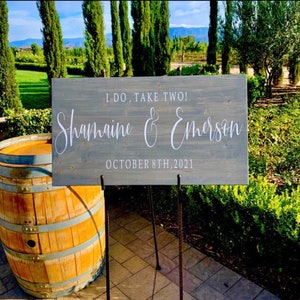 May include: A rustic wooden sign with the text "I do, take two!"  "Shamaine & Emerson" and "October 8th, 2021" in white lettering. The sign is standing on a black metal easel.