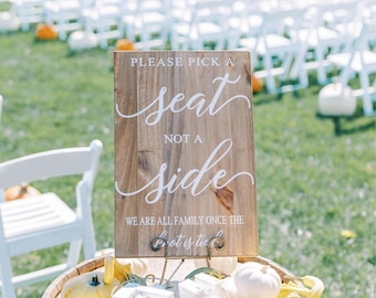 May include: A wooden sign with white text that reads "Please pick a seat, not a side. We are all family once the knot is tied." The sign is propped up on a barrel with white and yellow gourds and a wire basket filled with small white boxes.