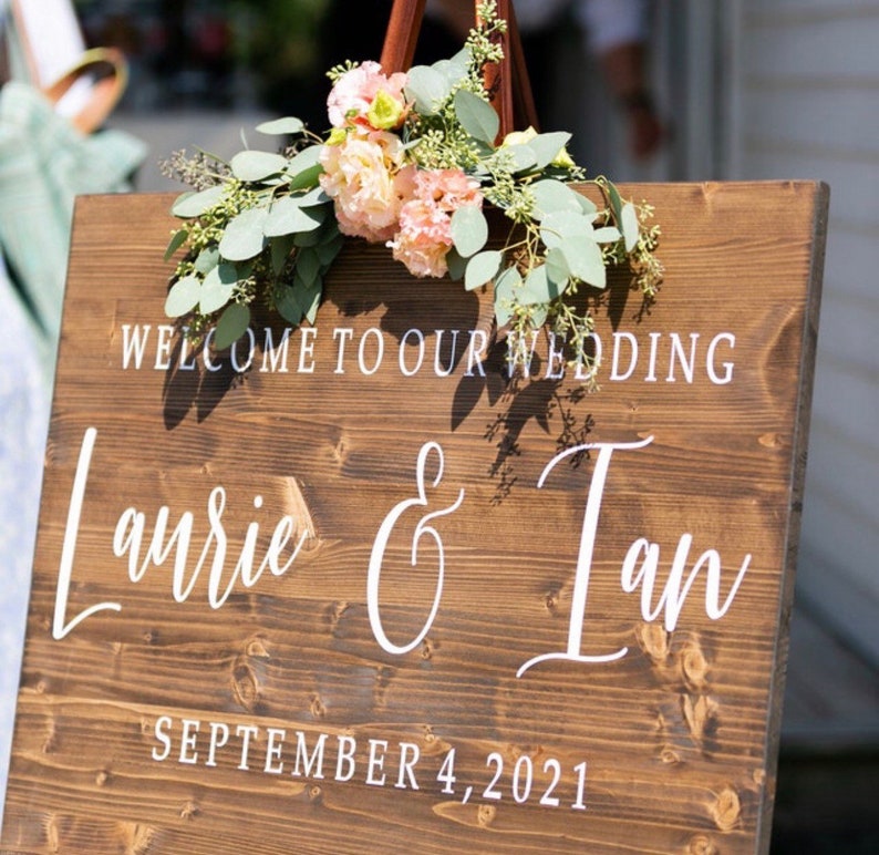 May include: A rustic wooden wedding sign with white text that reads "Welcome to our wedding Laurie & Ian September 4, 2021". The sign is decorated with a floral arrangement of pink and white flowers and green leaves.
