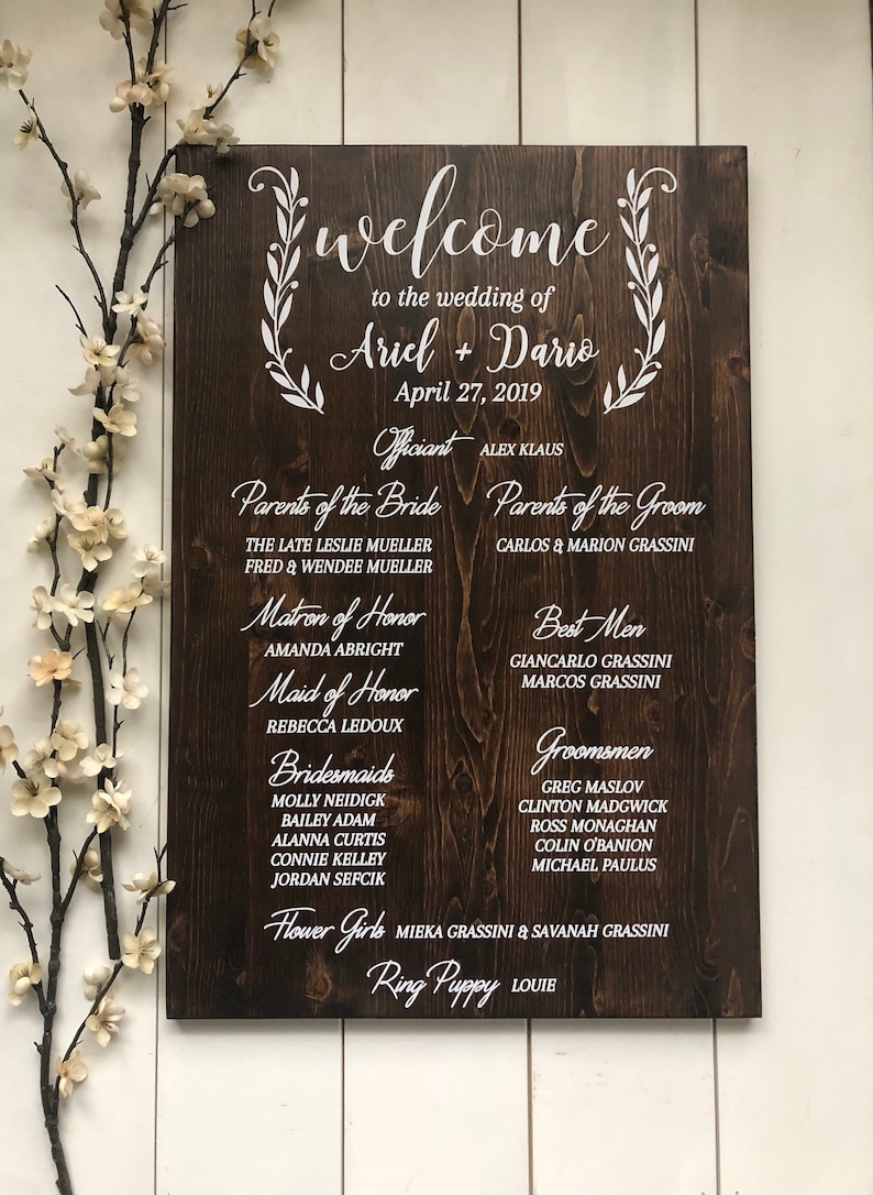 Puede incluir: Un letrero de madera r&uacute;stico con texto blanco anunciando una boda. El letrero incluye los nombres Ariel y Dario, la fecha de la boda, el nombre del oficiante y los nombres de los miembros de la fiesta de bodas.