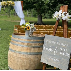 May include: A wooden sign with the text "Welcome to our wedding Kendra & TeRon June 26, 2021" on a wooden easel with white flowers. A wooden barrel sits in front of the sign.