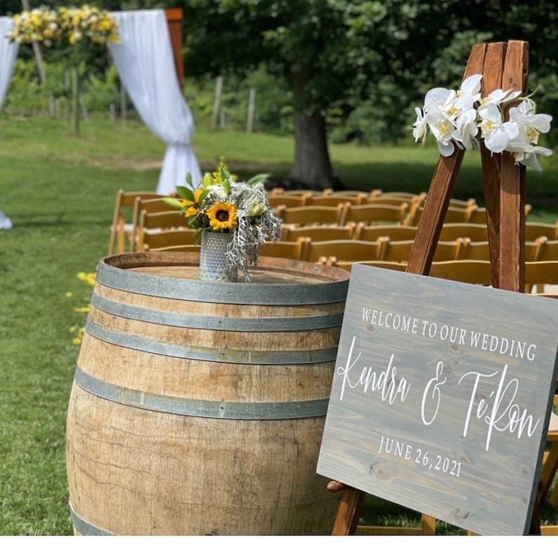 May include: A wooden sign with the text "Welcome to our wedding Kendra & TeRon June 26, 2021" on a wooden easel with white flowers. A wooden barrel sits in front of the sign.