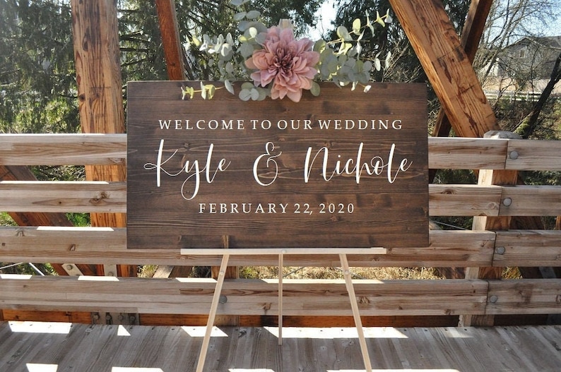 May include: A wooden wedding sign with the text "Welcome to our wedding Kyle & Nichole February 22, 2020" in white lettering. The sign is decorated with a pink flower and greenery.