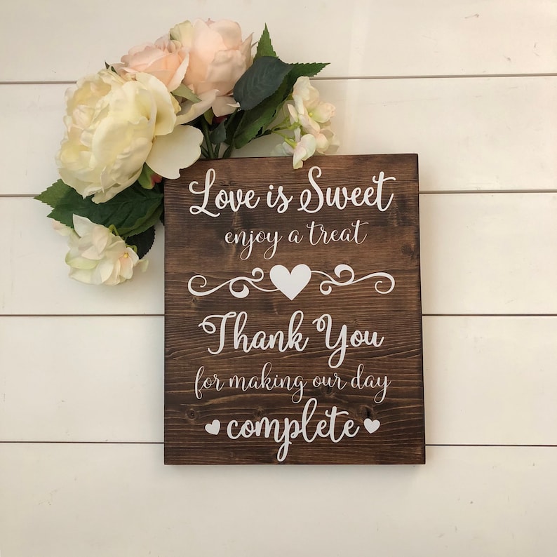 May include: A brown wooden sign with white text that reads "Love is Sweet enjoy a treat Thank You for making our day complete". The sign has a heart shape and a decorative swirl design.