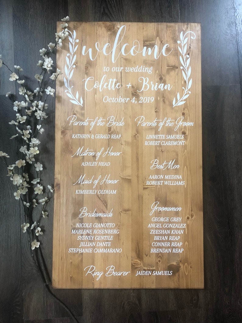 Puede incluir: Un letrero de madera con texto blanco anunciando una boda para Colette y Brian el 4 de octubre de 2019. El letrero enumera los nombres de los padres de la novia y el novio, la madrina de honor, la dama de honor, las damas de honor, los padrinos de boda y los padrinos de boda.
