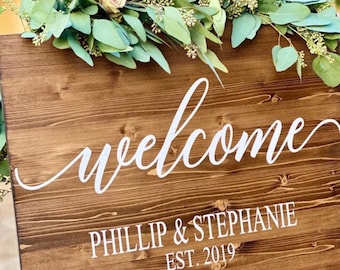 May include: A rustic wooden welcome sign with white lettering. The sign says "Welcome" in a cursive font and "Phillip & Stephanie Est. 2019" in a block font.