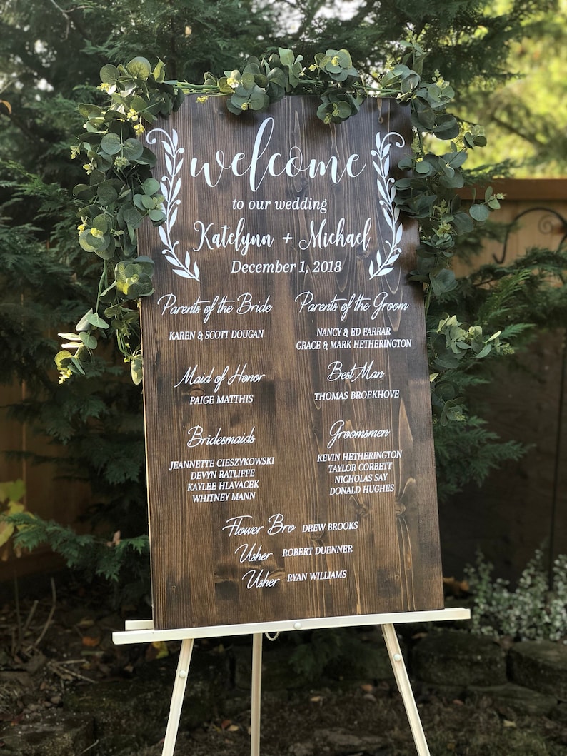 Puede incluir: Un cartel de bienvenida de boda de madera r&uacute;stica con caligraf&iacute;a blanca. El cartel enumera los nombres de la novia y el novio, Katelynn y Michael, la fecha de la boda, 1 de diciembre de 2018, y los nombres de los miembros de la fiesta de bodas.