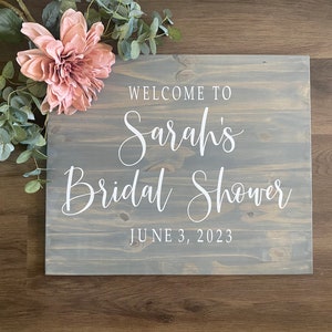 May include: A wooden sign with the text "WELCOME TO Sarah's Bridal Shower JUNE 3, 2023" in white script. The sign is gray and decorated with a pink flower and green leaves.