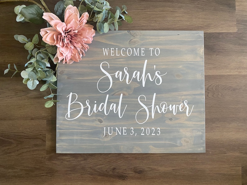 May include: A wooden sign with the text "WELCOME TO Sarah's Bridal Shower JUNE 3, 2023" in white script. The sign is gray and decorated with a pink flower and green leaves.