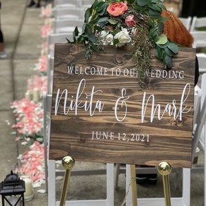 May include: A wooden wedding sign with white text on a gold easel. The sign reads "Welcome to our wedding Nikita & Mark June 12, 2021".