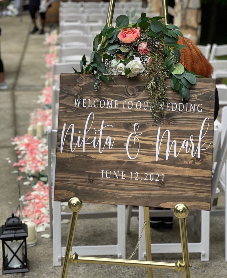 May include: A wooden wedding sign with white text on a gold easel. The sign reads "Welcome to our wedding Nikita & Mark June 12, 2021".
