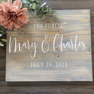 May include: A rustic wooden sign with a gray wash finish. The sign has white lettering that reads "The Furias Mary & Charles July 24, 2023".