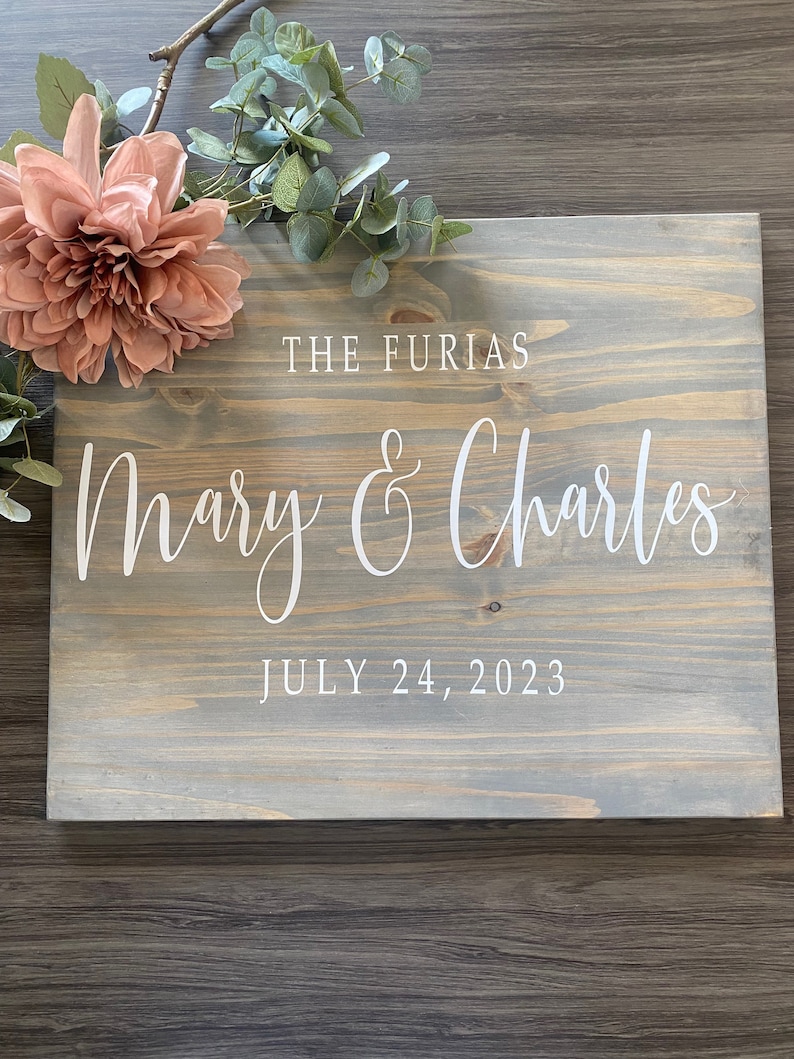 May include: A rustic wooden sign with a gray wash finish. The sign has white lettering that reads "The Furias Mary & Charles July 24, 2023".