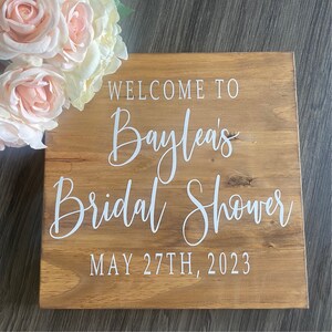 May include: A wooden sign with the text "WELCOME TO Baylea's Bridal Shower MAY 27TH, 2023" in white script. The sign is made of light brown wood and is decorated with light pink and white roses.