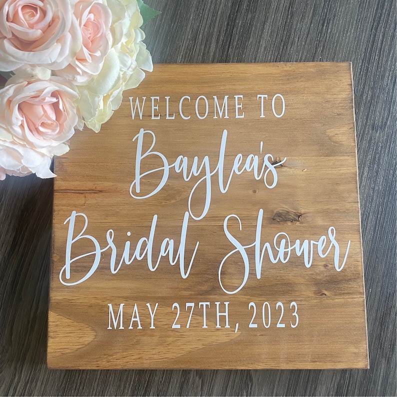 May include: A wooden sign with the text "WELCOME TO Baylea's Bridal Shower MAY 27TH, 2023" in white script. The sign is made of light brown wood and is decorated with light pink and white roses.