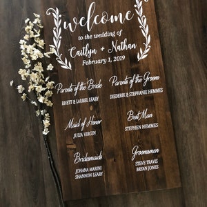 Puede incluir: Un letrero de madera con texto blanco anunciando una boda. El letrero dice "Bienvenido a la boda de Caitlyn + Nathan 1 de febrero de 2019". Tambi&eacute;n enumera los nombres de los padres de la novia y el novio, la dama de honor, las damas de honor, el padrino de boda y los padrinos de boda.