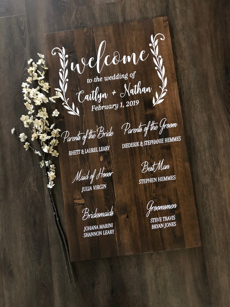 Puede incluir: Un letrero de madera con texto blanco anunciando una boda. El letrero dice "Bienvenido a la boda de Caitlyn + Nathan 1 de febrero de 2019". Tambi&eacute;n enumera los nombres de los padres de la novia y el novio, la dama de honor, las damas de honor, el padrino de boda y los padrinos de boda.