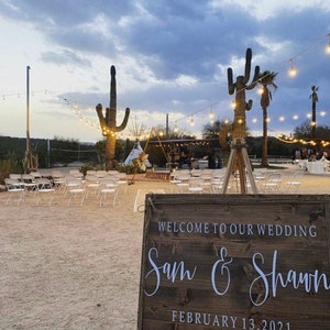 May include: A rustic wooden sign with white text announcing a wedding for Sam & Shawn on February 13, 2021. The sign is propped up on a tripod in front of a desert wedding ceremony setup with white chairs and string lights.