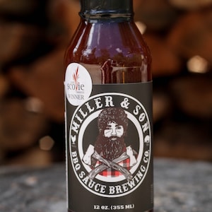May include: A 355 ml bottle of Miller & Son BBQ Sauce Brewing Co. sauce. The bottle has a black label with a white illustration of a lumberjack holding an axe. The label also includes the text "Scovie Awards Winner".