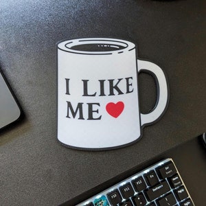May include: White coffee mug with black text that reads "I LIKE ME" and a red heart. The mug is on a black surface.