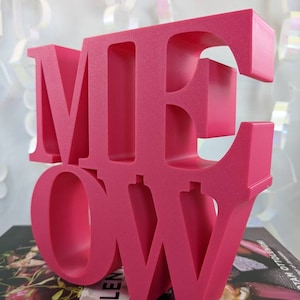 May include: A pink, 3D, freestanding sign that spells out the word "MEOW". The letters are stacked on top of each other, with the "M" on top and the "W" on the bottom.