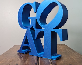 May include: A blue, three-dimensional word sculpture spelling out "GOAT". The letters are stacked on top of each other, with the "G" on top and the "T" on the bottom. The sculpture is sitting on a wooden surface.