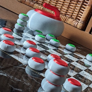 Könnte beinhalten: Nahaufnahme eines Schachbretts mit roten und grünen Spielsteinen. Das Schachbrett besteht aus schwarzen und weißen Feldern. Im Hintergrund befindet sich ein grauer Curling-Stein mit einem roten Griff.