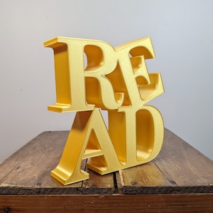 May include: A gold-colored, three-dimensional "READ" sign made of plastic. The letters are stacked on top of each other and are slightly angled.