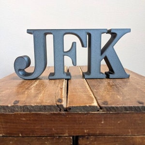 May include: Black wooden letters spelling out "JFK" on a wooden surface.