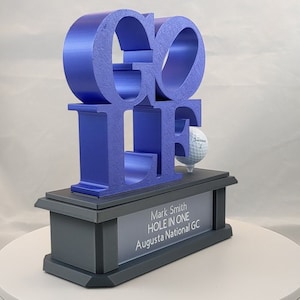 Puede incluir: Un trofeo de plástico azul con la palabra "GOLF" en letras grandes. El trofeo tiene una bola de golf en la parte superior y una placa que dice "Mark Smith HOLE IN ONE Augusta National GC".