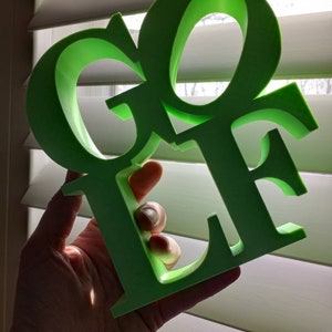 May include: A green 3D printed word "GOLF" with a stylised font. The word is held in a hand against a white window blind background.