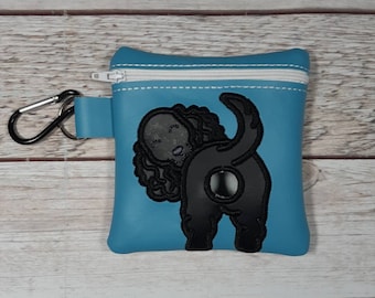Op de afbeelding: Een kleine blauwe ritszak met een zwart geborduurd hondenontwerp. De hond kijkt weg van de kijker en heeft een gekrulde staart.