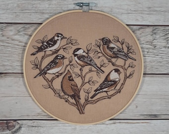 Op de afbeelding: Borduurwerk in een hoepel met een hartvormige opstelling van zes vogels op takken. De vogels zijn bruin, zwart en wit met gedetailleerde steken. De achtergrond is een licht beige stof.