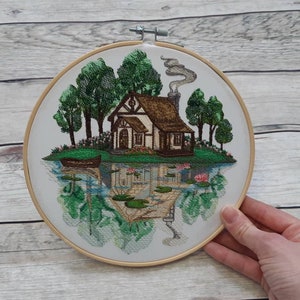 May include: Embroidered hoop art featuring a cottage with a brown roof and smoke coming from the chimney. The cottage is surrounded by green trees and a small boat on a lake with lily pads.