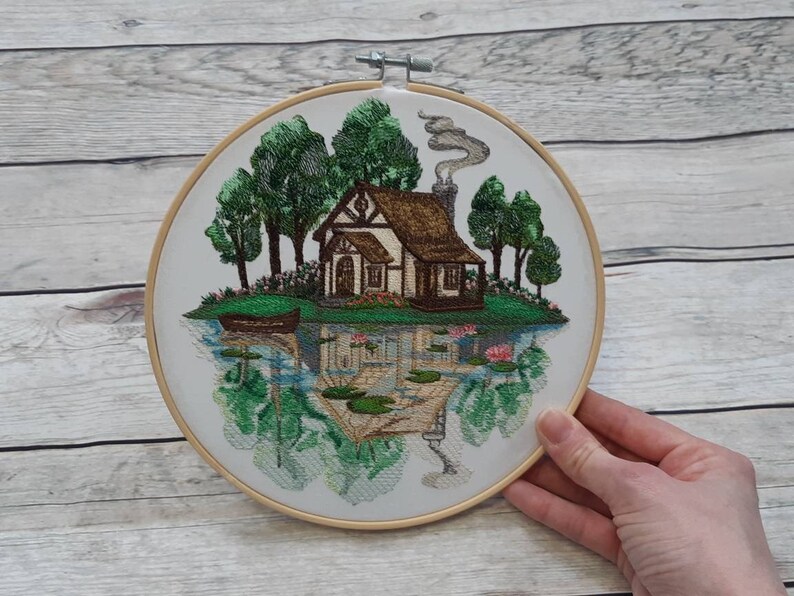 May include: Embroidered hoop art featuring a cottage with a brown roof and smoke coming from the chimney. The cottage is surrounded by green trees and a small boat on a lake with lily pads.