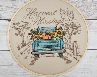 Op de afbeelding: Gestikte hoepelkunst met een blauwe pick-up truck die pompoenen en zonnebloemen vervoert. De truck rijdt over een landweg met bomen aan weerszijden. De tekst "Harvest Blessings" is boven de truck geborduurd.