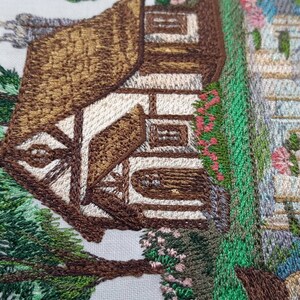 May include: Embroidered image of a brown cottage with a white door and windows, surrounded by green trees and flowers. The cottage is set against a white background.