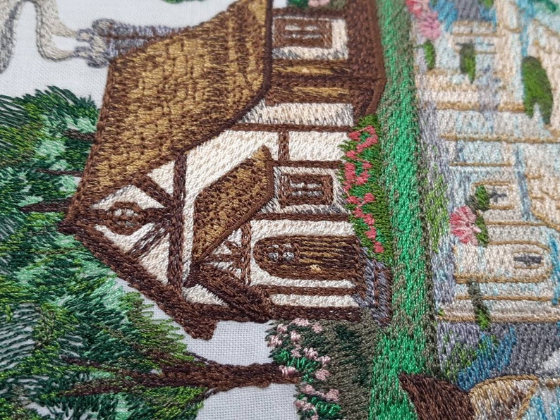 May include: Embroidered image of a brown cottage with a white door and windows, surrounded by green trees and flowers. The cottage is set against a white background.