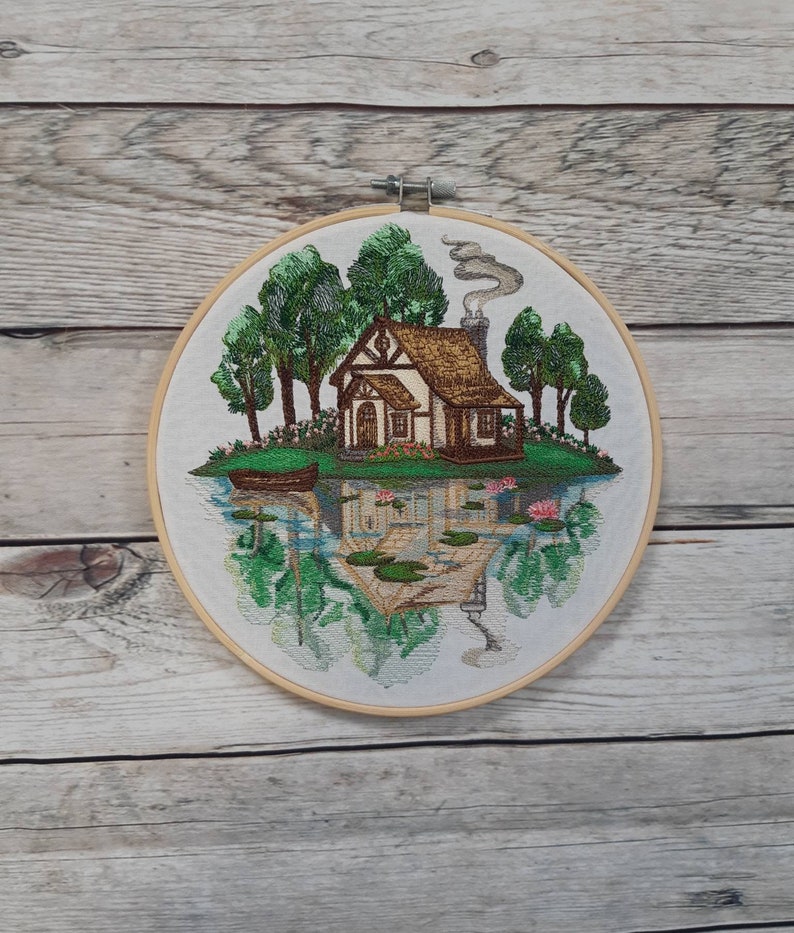 May include: Embroidered hoop art depicting a cottage with a brown roof and white walls, surrounded by green trees and a lake with lily pads and a small boat. The scene is reflected in the water.