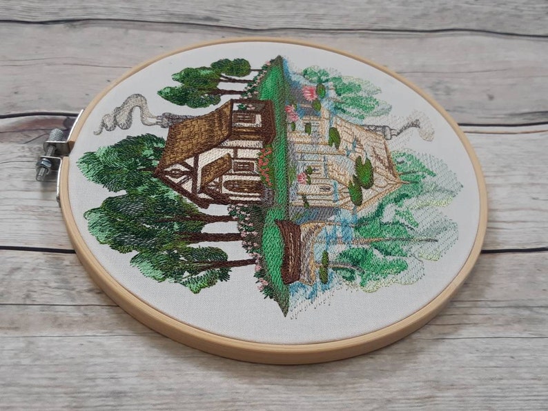 May include: Embroidered hoop art featuring a cottage with a brown roof and white walls surrounded by green trees. The cottage has a chimney with smoke coming out of it. The background is a light blue sky with white clouds.