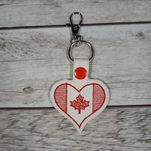 May include: A white heart-shaped keyring with a red embroidered Canadian flag. The keyring has a silver metal ring and a silver clip.